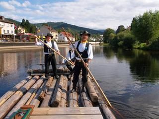 Zwei Flößer in der Tracht mit schwarzen Hosen, hohen schwarzen Stiefeln, einer schwarzen Weste und weißem Hemd auf dem Floß in der Murg