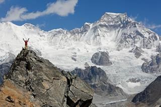 Eine Person steht im Bergpanorama auf einem Gipfel und reckt die Arme in die Höhe.
