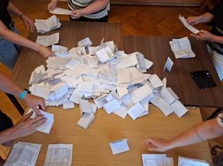 Hände von 5 Personen um einen Tisch beim Entfalten der Stimmzettel