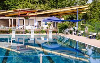 Auch in dieser Saison ist der Kiosk geöffnet. Schwimmbecken im Vordergrund. In der Mitte der Kiosk und im Hintergrund Bäume.