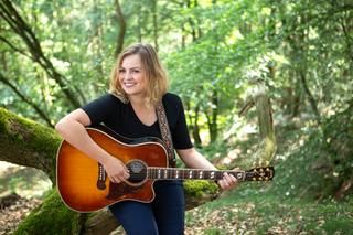 Eine junge Frau mit einer Gitarre lehnt im Wald an einem Baum. 