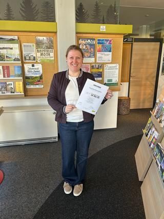 Kulturamtsleiterin Mußler freut sich über die Nachzertifizierung Eine Dame hält ein Zertifikat, dahinter ein Broschürenständer