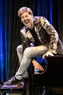 Daniel Helfrich Ein Kabarettist sitzt lachend auf einem Klavier.