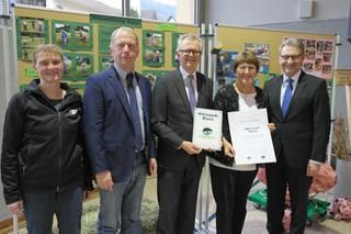Bürgermeister Dieter Knittel und Schulleiterin Brigitte Seiser halten die überreichte Auszeichnung in Händen. Sie stehen vor einer Bilderwand.  Auf dem Bild sind außerdem zwei Personen vom Naturpark und der Landrat.