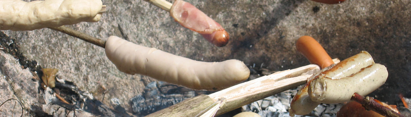 verschiedene Würste an Stecken brutzeln über dem Lagerfeuer