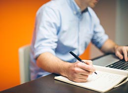 Foto: Pixabay.com Person sitz am Schreibtisch mit Laptol und Block und hat einen Stift in der Hand.