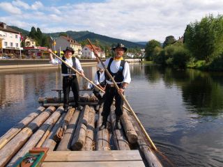 Zwei Flößer in der Tracht mit schwarzen Hosen, hohen schwarzen Stiefeln, einer schwarzen Weste und weißem Hemd auf dem Floß in der Murg