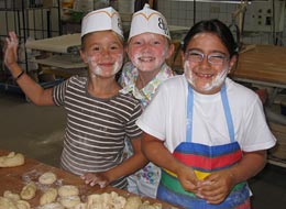Backen beim Kinderferienprogramm 3 Kinder mit mehlbestäubten Gesichtern in der Backstube formen Brötchen